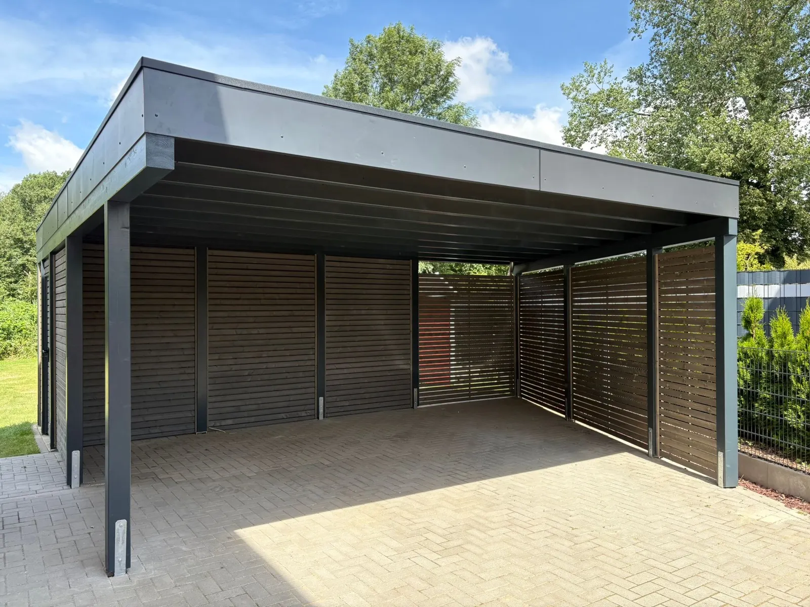 Doppelcarport mit anthrazitfarbenem Tragwerk und dunklen Holz-Lamellenwänden auf gepflasterter Einfahrt