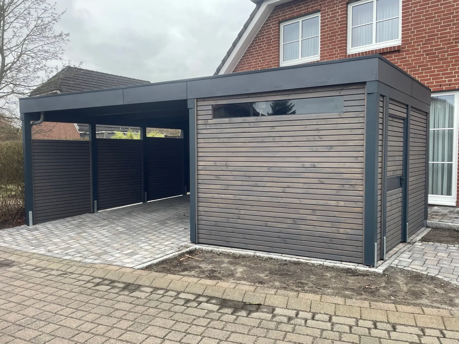 Doppelcarport aus grau lasiertem Holz mit integriertem Geräteraum und Flachdach an einem Klinkerhaus