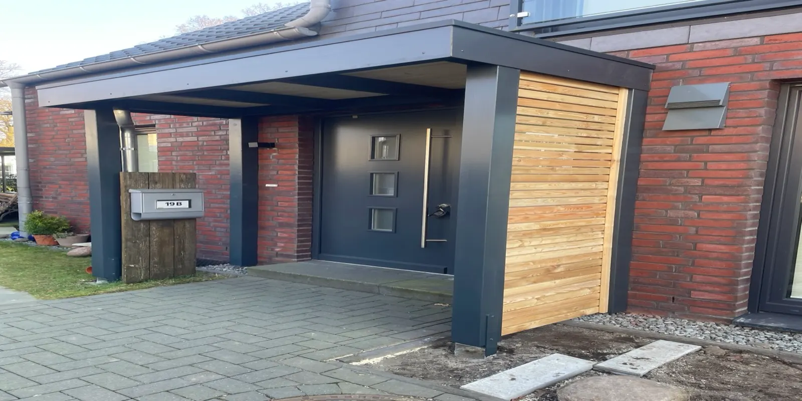 Anthrazitfarbenes Vordach mit Seitenteil aus Lärchenholz über einer Haustür an roter Klinkerfassade
